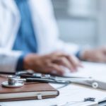 Uno stetoscopio e un taccuino su una scrivania in primo piano, con un medico sfocato sullo sfondo, che simboleggiano i fondi sanitari integrativi e il ruolo di Assidai