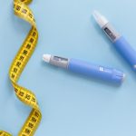 Due penne per l'iniezione di farmaci anti obesità accanto a un metro da sarto giallo su sfondo azzurro.