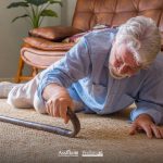 Un uomo anziano a terra dopo una caduta in casa, mentre cerca di raggiungere il suo bastone da passeggio.